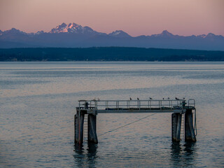 The setting sun casts the Cascade Mountains with pink as a standalone pier in Langley, with shore...