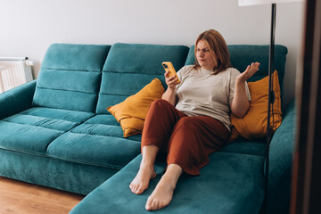 Obraz premium Upset sad, skeptical, serious woman holding smartphone gadget for communicate indoors. Negative people emotion, pensive girl sitting at home. Redhead woman holding smartphone. Screaming, hate, rage