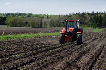 Obraz premium Red tractor plowing a freshly tilled field, with a backdrop of lush green trees under a clear sky