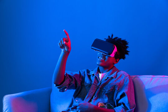 Young African American looking VR enjoy watching fantastic 3D pointing interesting object technology blue neon light virtual reality metaverse new world at living room sitting on sofa. Contrivance.