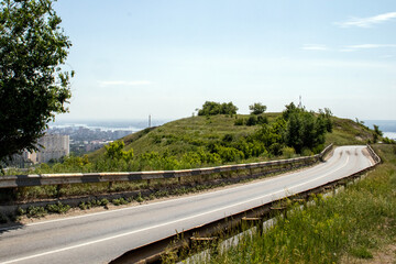The asphalt road goes around the hill to the horizon. The road around the hill among the green grass on a summer day. The road goes to the city on the horizon.