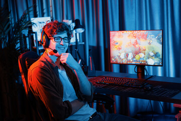 Host channel of young gaming streamer looking at camera to present fighting Moba battle arena game with multiplays team against on blurred pc screen background at neon digital lighting room. Gusher.