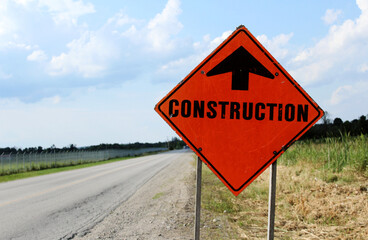 Orange construction sign with arrow pointing ahead at side of a rural road.
