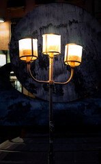 An old metal lantern with three lighted lamps casts a warm, inviting glow on the street at night.