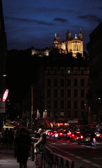 Obraz premium Lyon, France - August 12 2023. People stroll along the lively streets at night, illuminated by lanterns and shop signs, with the Basilica Notre Dame de Fourvière standing majestically in the backgroun