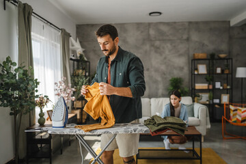 Adult man iron the wardrobe on the iron board while woman clean dust