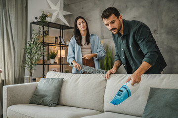 Obraz premium adult man vacuum sofa with hand vacuum cleaner while woman support him