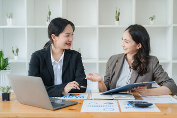 Strategic Collaboration: Two young Asian businesswomen engage in lively discussion over financial reports, their smiles reflecting a productive and positive work environment. 