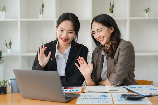 Virtual Collaboration, Real Connections: Two Asian businesswomen share a warm moment during an online meeting, waving at the screen with smiles that radiate the power of digital teamwork.