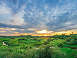 A stunning countryside sunset with a river winding through green meadows, painted skies of orange, pink, and blue, with fluffy clouds drifting by