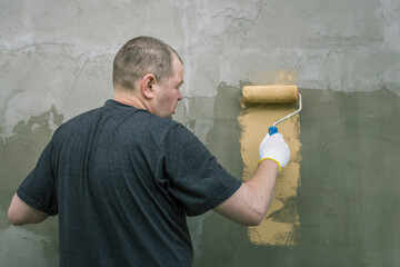 A man meticulously paints a wall with a roller, showing his dedication. The rough wall texture...