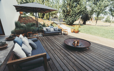 Outdoor patio area with wooden deck, cushioned sofas, and a fire pit. An umbrella provides shade,...