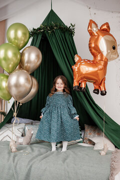 Child birthday party celebration. Happy little girl 4-5 years in stylish rustic style dress stands on bed decorated with baldachin and colorful foil balloons, smiles and looks at camera at bedroom