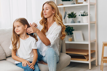 Mother lovingly combs daughter's hair as they sit together on a cozy living room couch