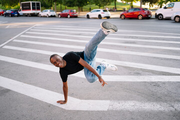 A young man in a black t-shirt and blue jeans performs a stunt in the middle of a street with white...