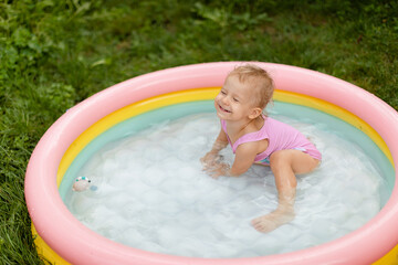 Laughing baby girl in a pink swimsuit is swimming in an inflatable pool in the backyard. 
