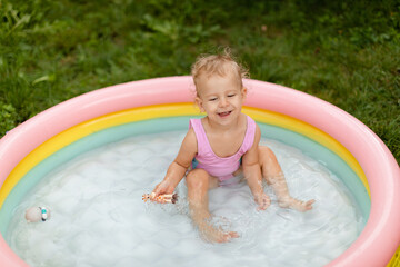 Laughing baby girl in a pink swimsuit is swimming and playing with toys in an inflatable pool in the backyard. Lifestyle. 