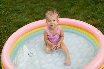 Laughing baby girl in a pink swimsuit is swimming in an inflatable pool in the backyard. 