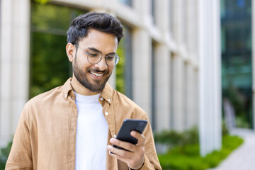 Man wearing glasses and casual clothing smiling while using smartphone outdoors near modern building. Person enjoying communication and technology in outdoor setting on sunny day