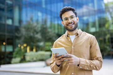 Smiling young man using digital tablet in modern urban environment. Confident individual in casual outfit holding device outdoors. Concept of technology, business, and lifestyle in city setting.