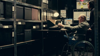 Detective with impairment develops case timelines and profiles using archival records and research, disability friendly archive room. Senior agent in wheelchair interpreting legal files. Camera B.