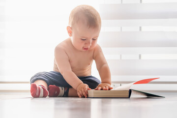 Naklejka premium Child development concept. Funny portrait of sweet baby boy sitting on the floor at home and reading book. Horizontal image.