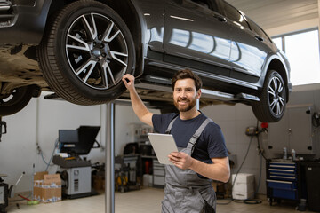 Smiling mechanic using tablet in auto repair shop with car on lift. Confident male technician...
