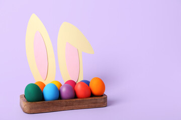 Box with Easter eggs and bunny ears on lilac background