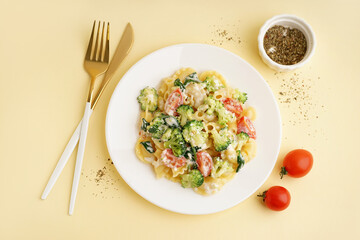 Plate of tasty pasta with broccoli, tomatoes, cutlery and spices on yellow background