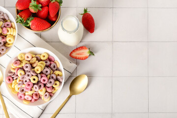 Bowls with colorful sweet cereal rings, strawberries and bottle of milk on white tile table