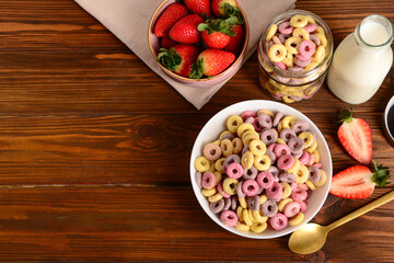 Bowls with colorful sweet cereal rings, strawberries and bottle of milk on wooden table