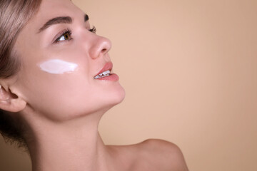 Fototapeta premium Beautiful woman with cream on her face against beige background, closeup. Space for text