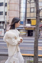 Twenty-something Chinese woman with black hair wearing a white coat with a fluffy fur hood and a white skirt walking on a public road in Minato, Tokyo, Japan.