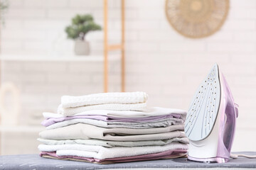 Iron and stack of clothes on board on blurred background