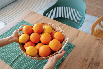 Female hands putting wicker basket with citruses on table in light kitchen