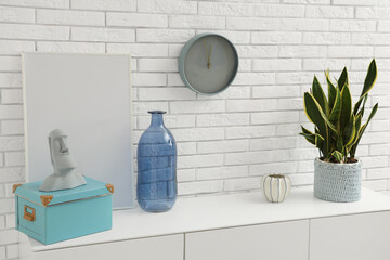 Decorative bottle, houseplant and statuette on modern cabinet near white brick wall