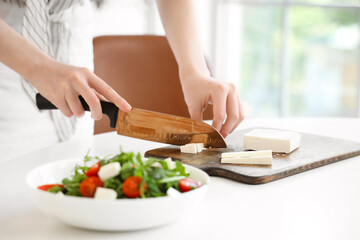 Young woman cutting feta cheese for salad in kitchen
