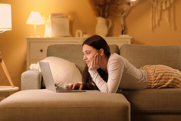 Obraz premium Young woman in pajamas lying on soft couch and chatting with modern laptop in living room at night