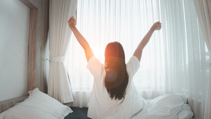 A young woman wakes up in a bright morning. Stretches in the Bed, Sun Shines on Her From the Big Window. Happy Young Girl Greets New Day with Warm Sunlight Flare.