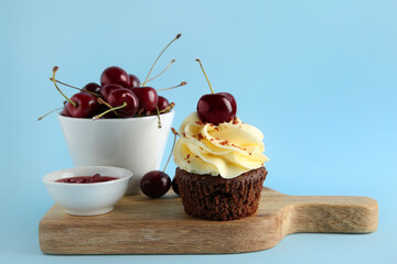 Wooden board with sweet cherry cupcake and bowl of jam on blue background