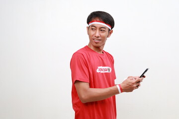 Asian young boy with red white ribbon for celebrating Indonesia independence day standing looking sideways while holding cell phone and Indonesian flag