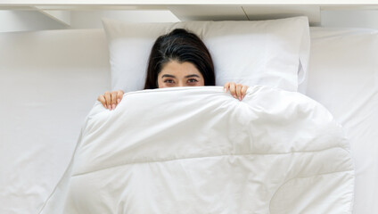 A woman is lying in bed, partly hidden under a white blanket, with a playful expression in her eye.