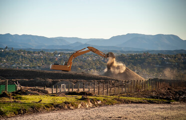 An excavator dumping na load of dirt creating dust backlit with mountains in the background