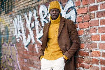Obraz premium Stylish African American man in yellow beanie and brown coat posing confidently against urban brick wall with graffiti, showcasing trendy winter street fashion