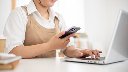 Fototapeta premium An Asian woman working remotely from a cafe, multitasking, using her smartphone and her laptop.
