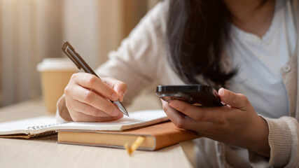 A woman's hand holding a smartphone and a pen, using her smartphone while taking notes in a notebook