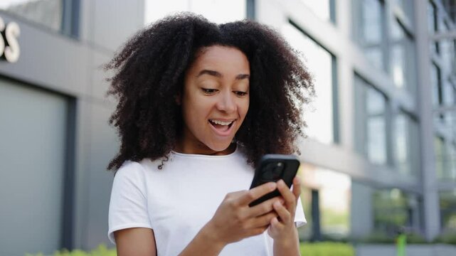 Attractive female in light clothes using smartphone while receiving message with good news from distant friend. Young brunette rejoicing while checking mailbox while having walk in business district.