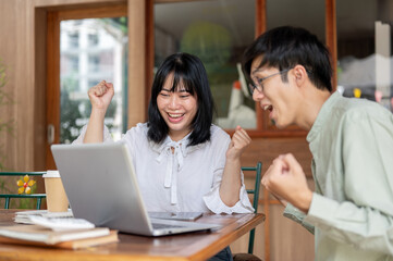 Fototapeta premium Two cheerful Asian friends, male and female, are sitting at a cafe, celebrating good news together.