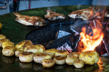 A fire burns inside a large round metal grill. Baked potatoes lie on the steel surface and flounder fish is fried. Background.