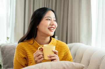 A beautiful Asian woman is holding a coffee mug, sitting on a couch, smiling and daydreaming.